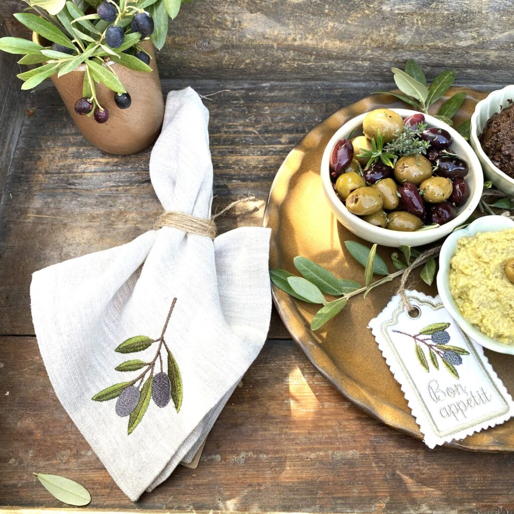 Handgefertigte mediterrane Serviette aus naturfarben meliertem Leinen mit gesticktem Olivenzweig von Atelier Luhabru, gefaltet auf rustikalem Holztisch